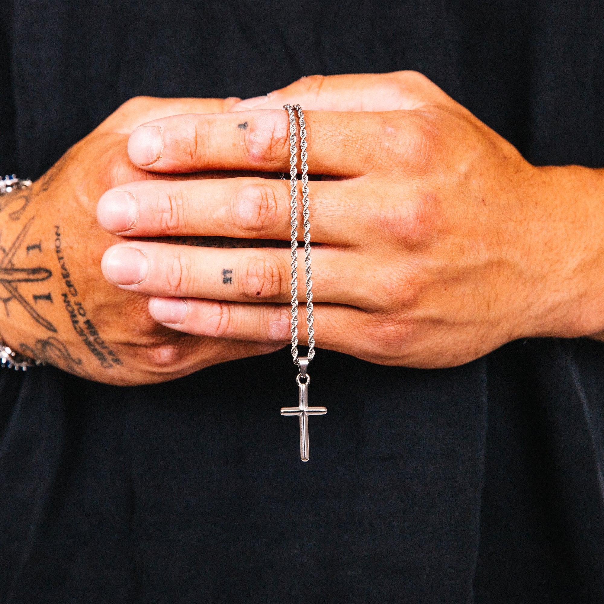 Silver cross necklace held between tattooed hands against a black background